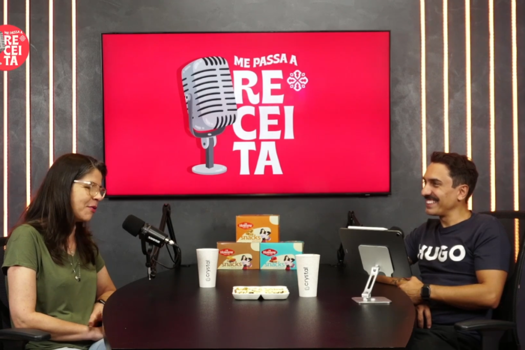 podcast episódio me passa a receita vinícius e marcia estão sentados frente à frente em uma mesa com uma teve ao fundo com o logo me passa a receita